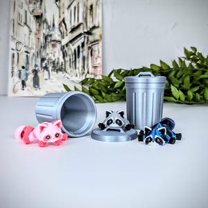 May include: Three small raccoon figurines, one pink, one gray, and one blue, are arranged around a silver trash can. The trash can is a miniature version of a standard trash can with a lid.
