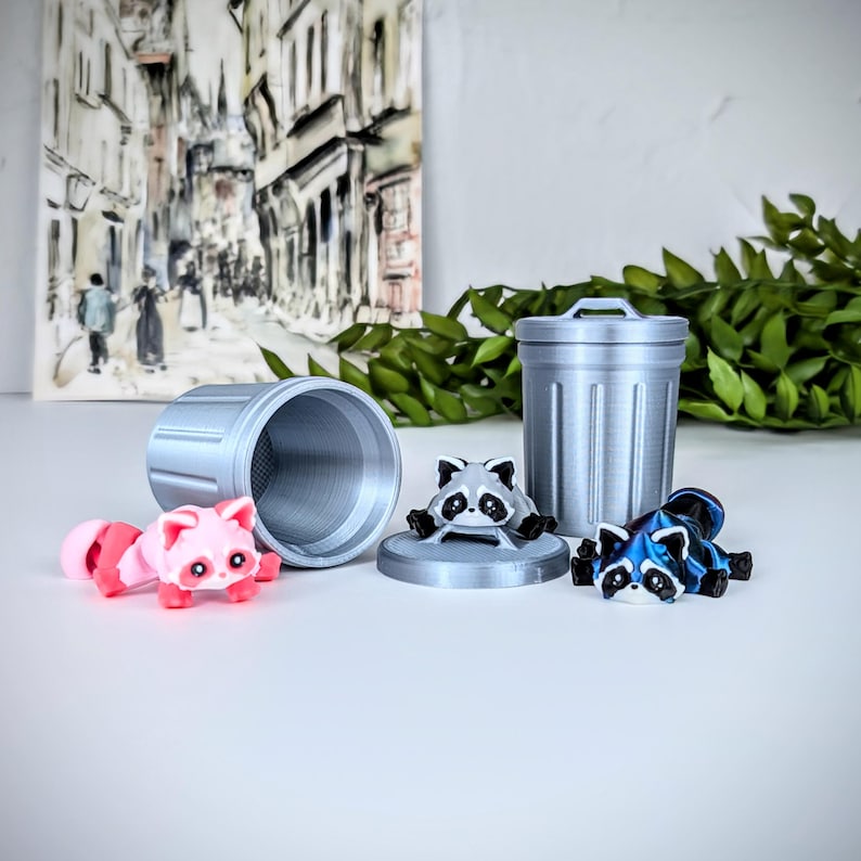 May include: Three small raccoon figurines, one pink, one gray, and one blue, are arranged around a silver trash can. The trash can is a miniature version of a standard trash can with a lid.