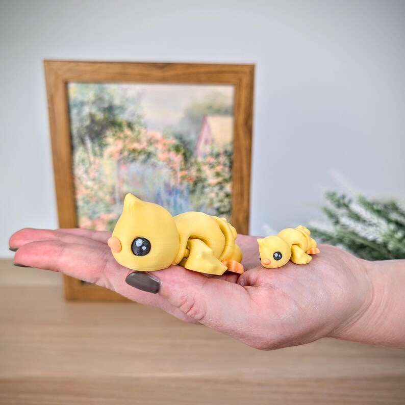 May include: Two yellow duck figurines, one large and one small, with black eyes and orange beaks. The larger duck is lying down, and the smaller duck is next to it. The figurines are on a hand, with a framed painting in the background.