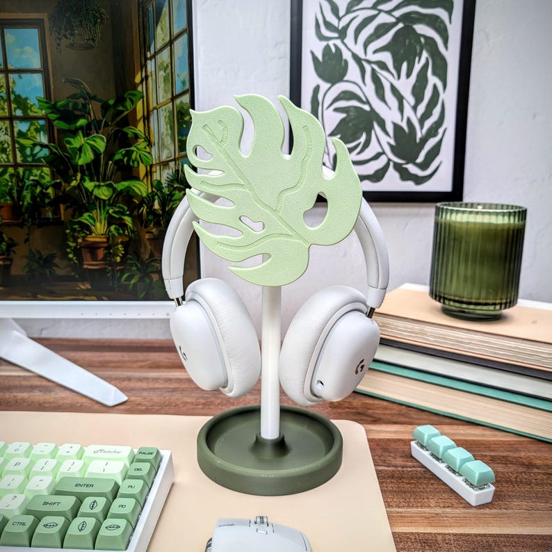 May include: A green monstera leaf shaped headphone stand with a white pair of headphones hanging on it. The stand is on a beige surface with a green keyboard and a white mouse.