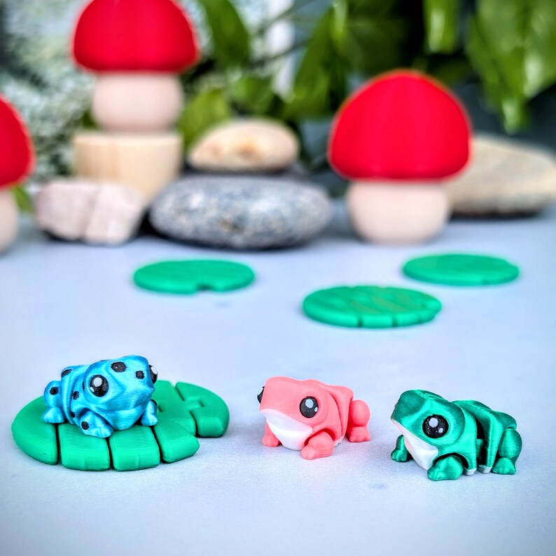 May include: A whimsical scene featuring miniature, colorful frog figurines on green lily pads. The frogs are in shades of blue, pink, and green. Red mushroom figurines and smooth stones are in the background, creating a playful, nature-inspired aesthetic.