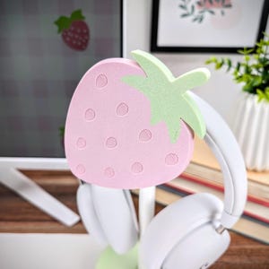 May include: A pink and green strawberry-shaped headphone stand. The stand is made of foam and has a textured surface. The stand is holding a pair of white headphones.