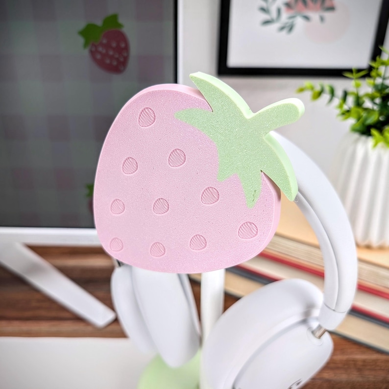 May include: A pink and green strawberry-shaped headphone stand. The stand is made of foam and has a textured surface. The stand is holding a pair of white headphones.