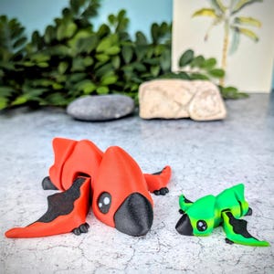 May include: Two small, hand-painted, ceramic figurines of mythical creatures. The larger figurine is red with black accents and has wings. The smaller figurine is green with black accents and has wings. Both figurines have large, black eyes.