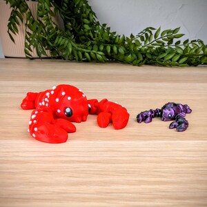 May include: A red lobster figurine with white spots and a purple scorpion figurine. Both figurines have black eyes and are posed on a wooden surface.