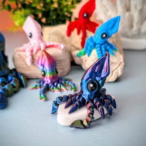May include: A collection of colorful 3D printed octopus figurines. The octopus figurines are in various colors, including pink, blue, red, and iridescent purple. The octopus figurines are sitting on a white surface.