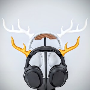 May include: A black over-ear headphone with gold antlers on a white deer head stand. The stand has a wooden base.