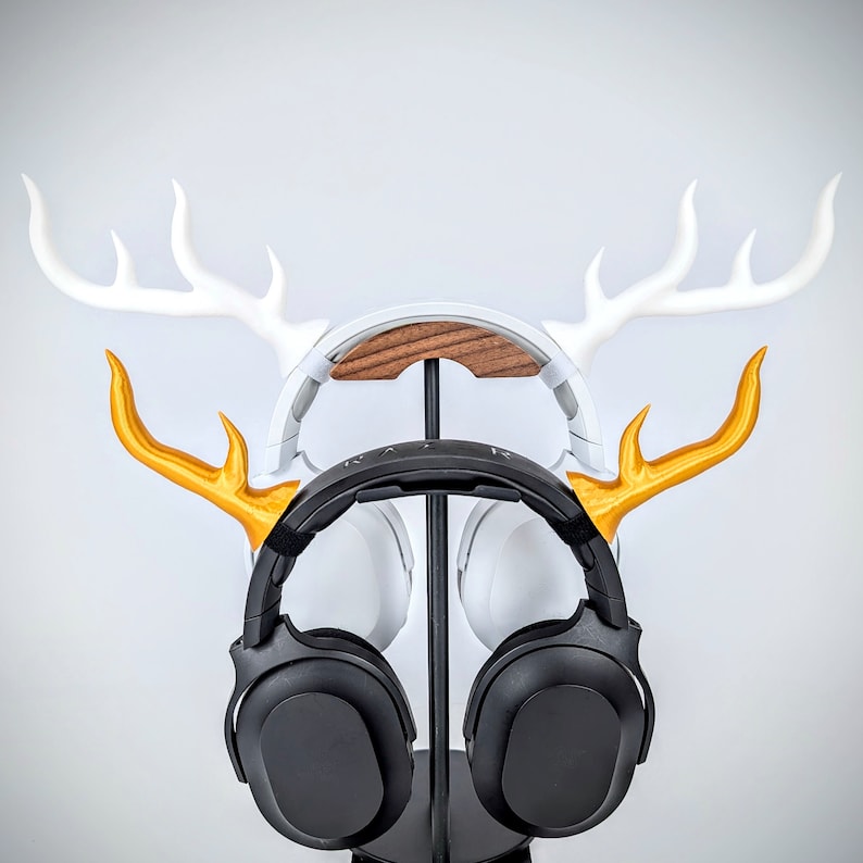 May include: A black over-ear headphone with gold antlers on a white deer head stand. The stand has a wooden base.