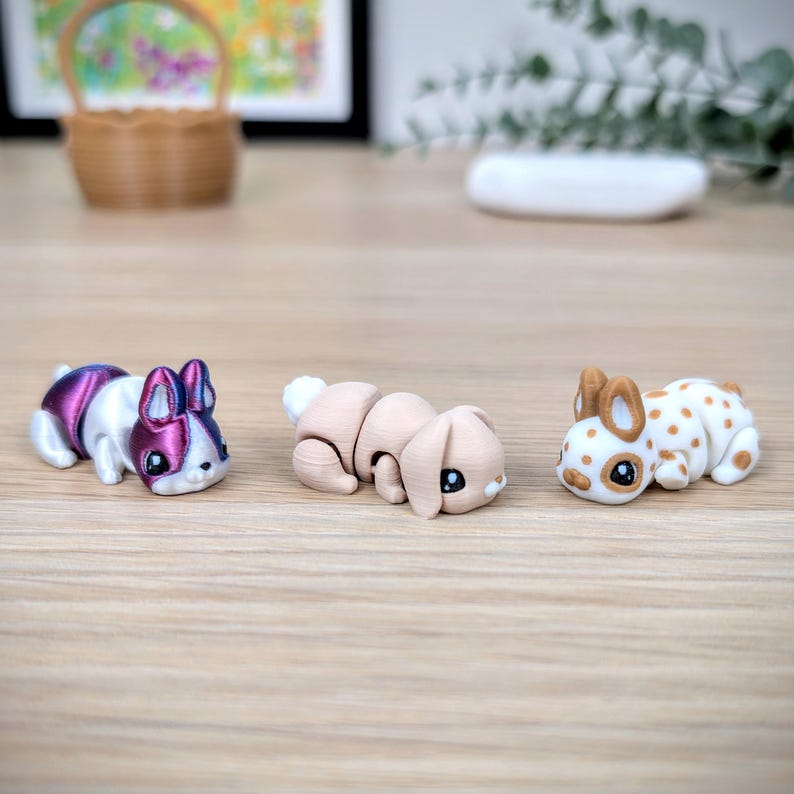 May include: Three small bunny figurines in different colours and patterns. One is white and purple, another is tan, and the third is white with brown spots. They are on a light brown wooden surface.