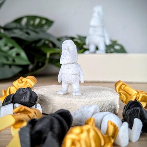 May include: A white, speckled figurine with a rounded head and short limbs stands on a white platform. The figurine is surrounded by a variety of small, colorful figurines.