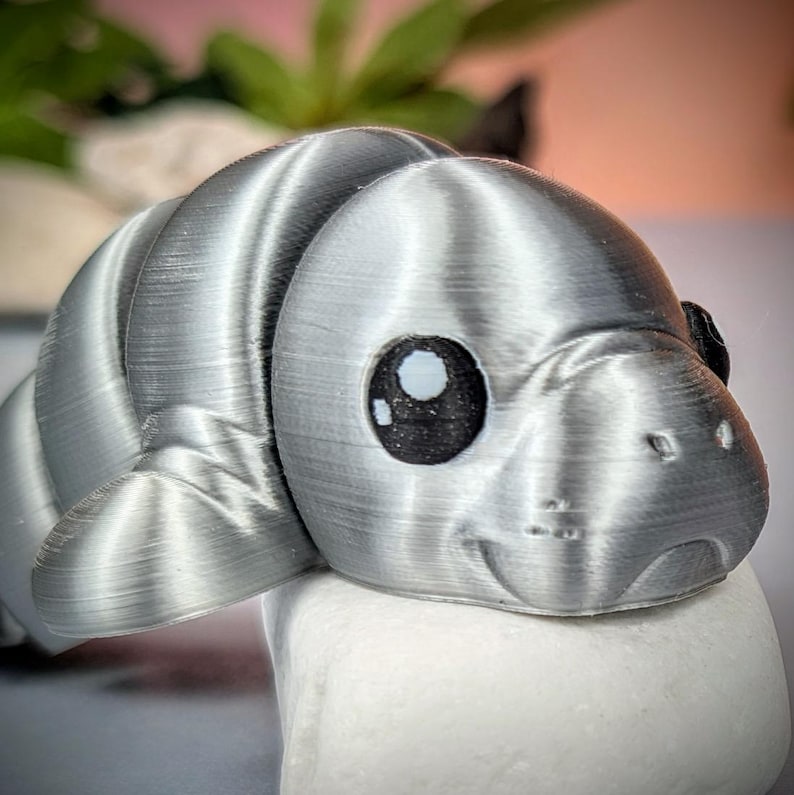 May include: A 3D-printed silver manatee figurine with large, expressive eyes and a smiling face. The manatee is resting on a white, rounded surface. The background is blurred with green and pink hues.