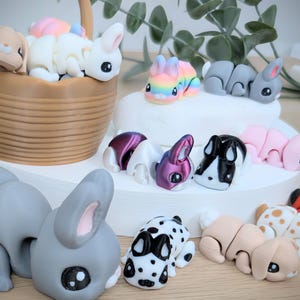 May include: A selection of miniature bunny figurines in various colours. The bunnies include white, grey, pink, and rainbow designs. Some are in a basket, others arranged on a white surface. The bunnies have large eyes and are in playful poses. A charming collection.