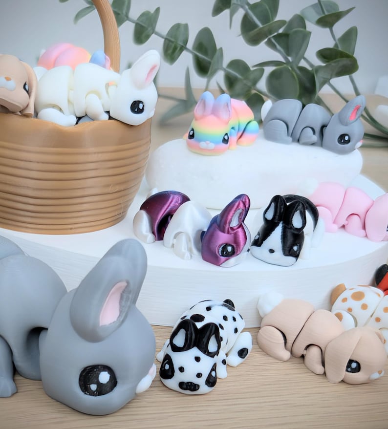 May include: A selection of miniature bunny figurines in various colours. The bunnies include white, grey, pink, and rainbow designs. Some are in a basket, others arranged on a white surface. The bunnies have large eyes and are in playful poses. A charming collection.
