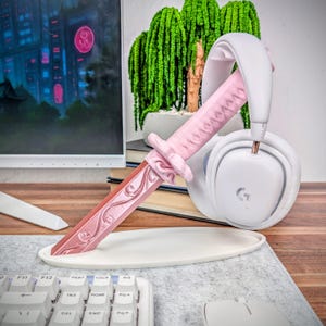 May include: A pink decorative katana rests on a white oval stand, next to a pair of white headphones. The katana features an ornate handle and blade design. A computer monitor and keyboard are in the foreground, with a small potted plant in the background.
