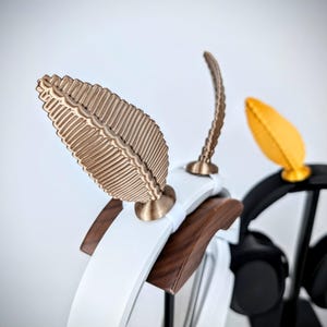 May include: A white and brown wooden headphone stand with two gold leaf-shaped hooks. The stand is designed to hold headphones.