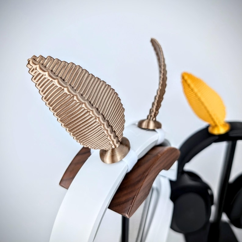May include: A white and brown wooden headphone stand with two gold leaf-shaped hooks. The stand is designed to hold headphones.