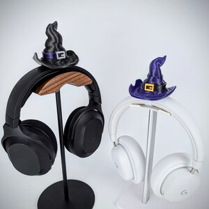 May include: Two black and white headphones with witch hats on stands. The left headphone has a black witch hat with a purple band and the right headphone has a purple witch hat with a yellow buckle.