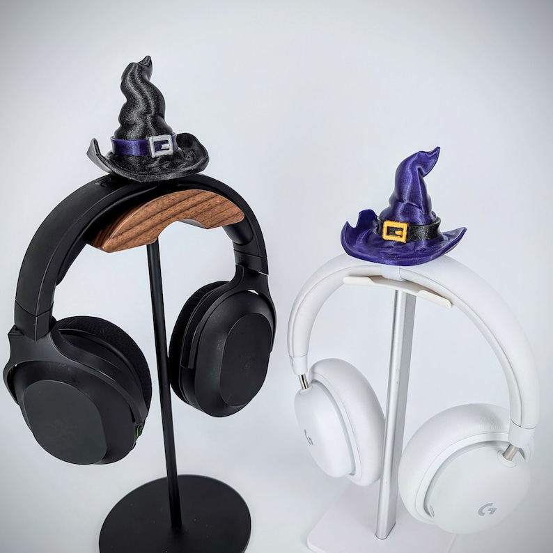 May include: Two black and white headphones with witch hats on stands. The left headphone has a black witch hat with a purple band and the right headphone has a purple witch hat with a yellow buckle.