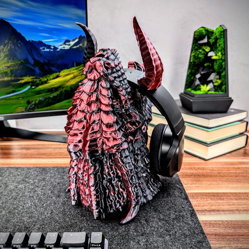 May include: A red and black dragon-shaped headphone stand with a pair of black headphones resting on the horns. The stand is made of a 3D printed material and has a detailed, textured surface.