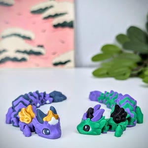 K&ouml;nnte beinhalten: Zwei bewegliche Drachenfiguren. Einer ist lila mit goldenen und blauen Akzenten, der andere ist gr&uuml;n mit lila und schwarzen Akzenten. Die Drachen befinden sich auf einer wei&szlig;en Oberfl&auml;che, mit einem rosa-wei&szlig;en Gem&auml;lde im Hintergrund.