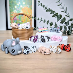 May include: A selection of miniature bunny figurines in various colours. Some are in a woven basket, others are on a white platform. The bunnies are grey, pink, black, white, and red. The overall theme is whimsical and decorative, suitable for collectors.