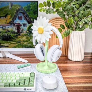 May include: A green and white daisy-shaped headphone stand with a pair of white headphones hanging on it. The stand is on a gray desk with a white keyboard and a white mouse.