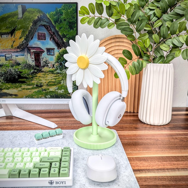 May include: A green and white daisy-shaped headphone stand with a pair of white headphones hanging on it. The stand is on a gray desk with a white keyboard and a white mouse.