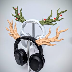 May include: Two pairs of headphones are displayed on a silver stand. One pair is black, the other white. Both have decorative antler-shaped attachments. The antlers are tan and green, with small red mushroom accents, creating a whimsical forest theme.