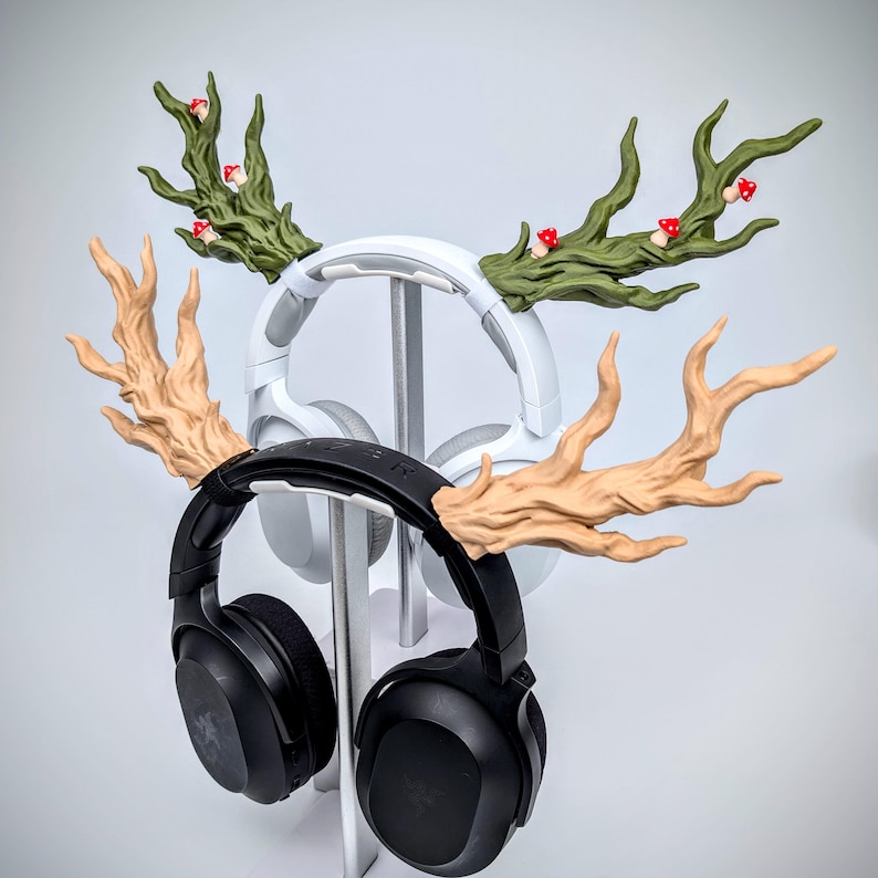May include: Two pairs of headphones are displayed on a silver stand. One pair is black, the other white. Both have decorative antler-shaped attachments. The antlers are tan and green, with small red mushroom accents, creating a whimsical forest theme.