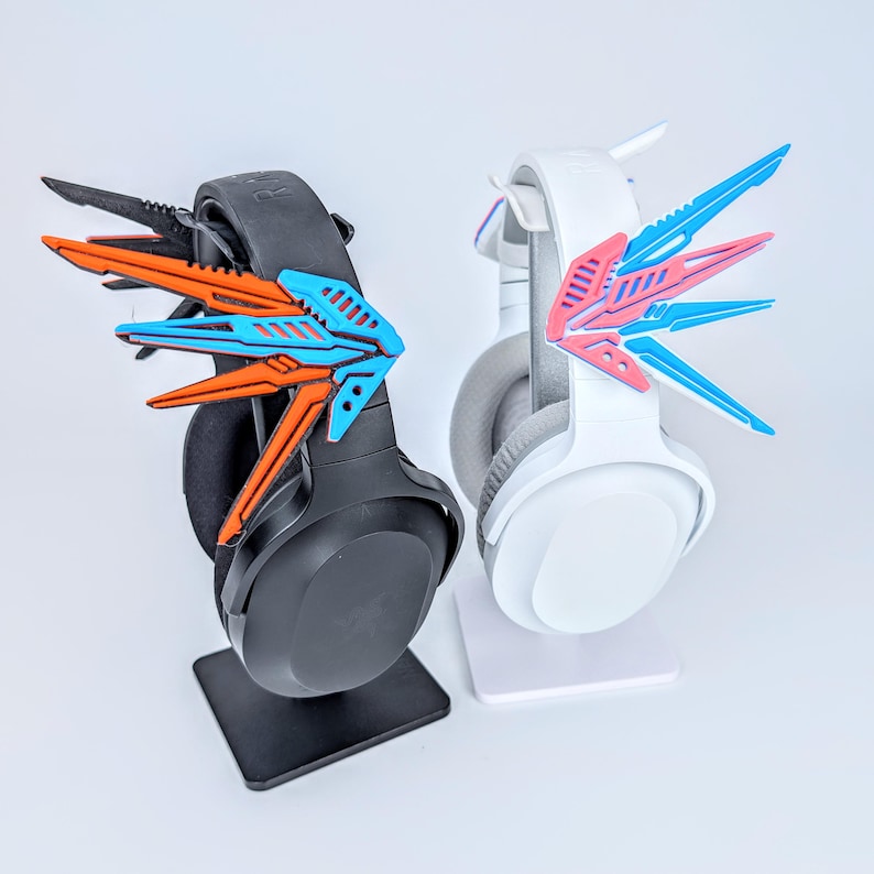 May include: Two gaming headsets, one black and one white, each adorned with colorful, stylized wing-shaped accessories. The black headset features orange and blue wings, while the white headset has pink and blue wings. Both are displayed on stands.
