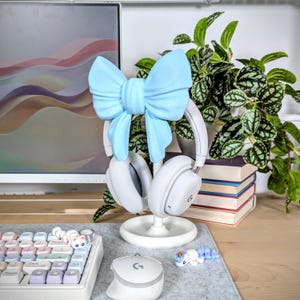 May include: A white headset rests on a white stand, adorned with a light blue bow. The setup includes a matching white mouse and keyboard with pastel-colored keys. A small, decorative unicorn figurine and a potted plant complete the scene.