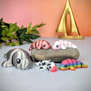 May include: A selection of colourful, handcrafted manatee figurines. The manatees are in various colours, including silver, pink, white with pink hearts, black and white, and rainbow. They are arranged on a grey surface with a gold sailboat in the background.