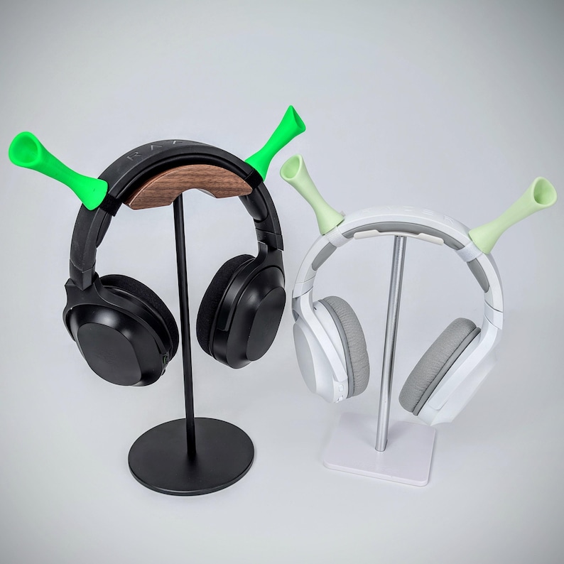 May include: Two headphone stands with green horn-shaped accents. One stand is black with a wooden headphone rest, holding black headphones. The other is white with silver accents, holding white and gray headphones. Both stands have a round base.