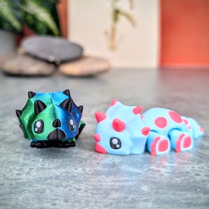 May include: Two small, 3D printed dinosaur figurines. One is green and blue with black horns and the other is light blue with pink spots and horns.