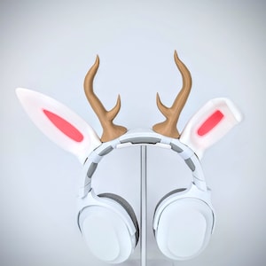 May include: White headphones with a white headband and large earcups. The headband has two white bunny ears with pink accents and two brown deer antlers.