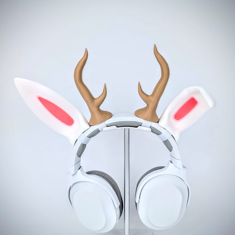 May include: White headphones with a white headband and large earcups. The headband has two white bunny ears with pink accents and two brown deer antlers.