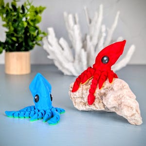 May include: Two colorful octopus figurines, one red and one blue, are displayed on a white rock. The red octopus is perched on top of the rock, while the blue octopus is lying on the surface.