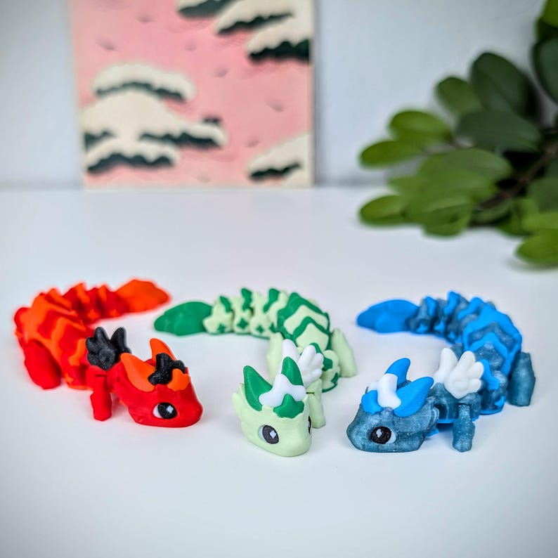 K&ouml;nnte beinhalten: Drei farbenfrohe Drachenfiguren. Einer ist rot und orange, ein anderer gr&uuml;n und wei&szlig;, und der dritte blau und wei&szlig;. Die Drachen stehen auf einer wei&szlig;en Oberfl&auml;che, mit einem rosa-wei&szlig;en abstrakten Gem&auml;lde im Hintergrund.