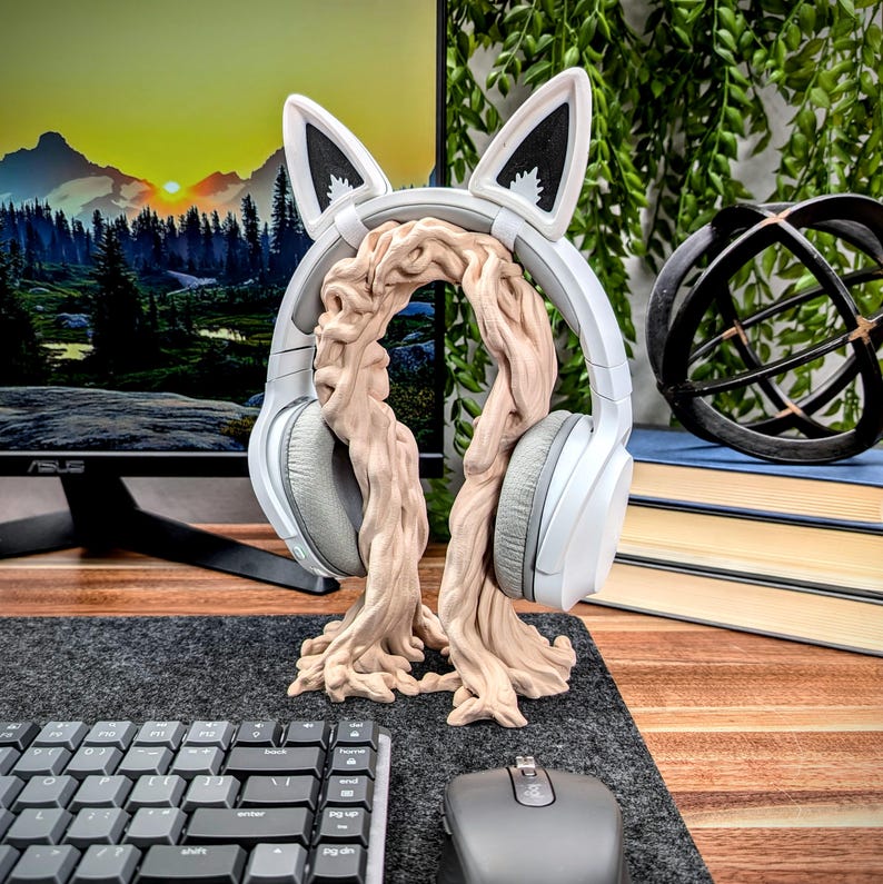 May include: White headphones with cat ears are resting on a wooden stand shaped like a tree trunk. The stand is on a gray desk mat with a keyboard and mouse.