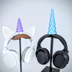 May include: Two pairs of headphones with unicorn-themed stands. One pair is white with purple ears and a horn, the other is black with a blue horn.