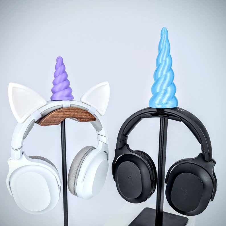 May include: Two pairs of headphones with unicorn-themed stands. One pair is white with purple ears and a horn, the other is black with a blue horn.