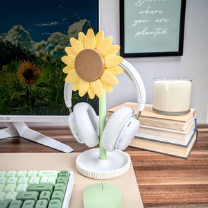 K&ouml;nnte beinhalten: Ein wei&szlig;er Kopfh&ouml;rerst&auml;nder mit Sonnenblumen-Design. Der St&auml;nder hat einen gr&uuml;nen Stiel und eine gelbe Sonnenblume mit brauner Mitte. Der St&auml;nder h&auml;lt ein Paar wei&szlig;e Kopfh&ouml;rer.