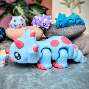 May include: A light blue articulated toy dinosaur with pink spots. The dinosaur is a triceratops with three horns on its head. The toy is posed with its head down and its tail curled up.