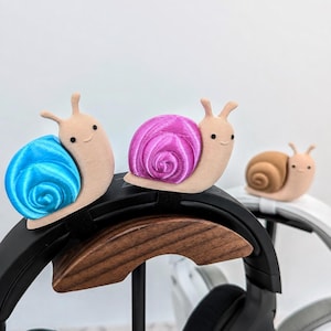 May include: Three small, beige snail-shaped cable organisers with smooth, rounded bodies and black dot eyes. Each snail features a vibrant, spiraled shell in bright blue, magenta, or tan, designed to clip onto the headband of black or white headphones.