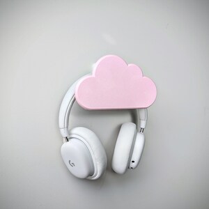 May include: White headphones hanging on a pink cloud-shaped headphone holder mounted on a white wall.