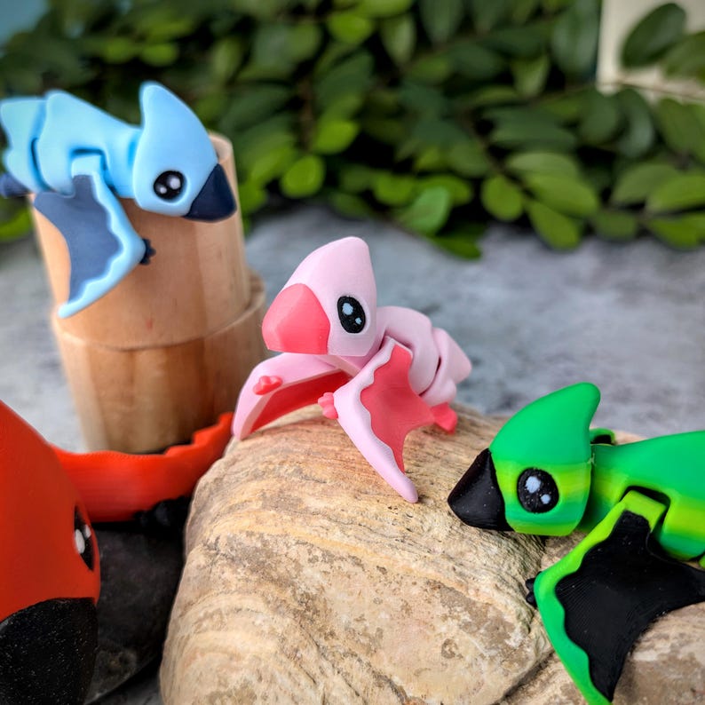 May include: Three small, colorful, articulated toy dinosaurs. The dinosaurs are posed with their wings spread out. The dinosaurs are blue, pink, and green with black accents.