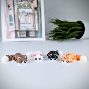 May include: Four toy cat figures in brown, white, black, and orange with white tails. The cat toys are lying down on a white surface. The background includes a framed picture and a white pot with green plants.