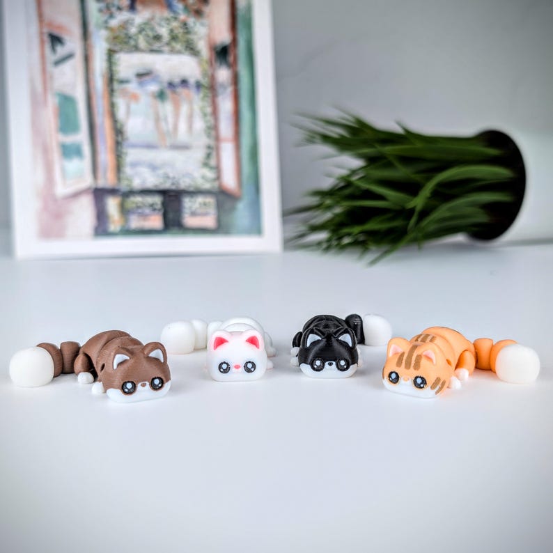 May include: Four toy cat figures in brown, white, black, and orange with white tails. The cat toys are lying down on a white surface. The background includes a framed picture and a white pot with green plants.
