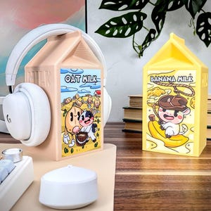 May include: Two cartoon milk carton shaped headphone stands. The oat milk carton is beige with a cartoon oat and cow. The banana milk carton is yellow with a cartoon cow riding a banana.