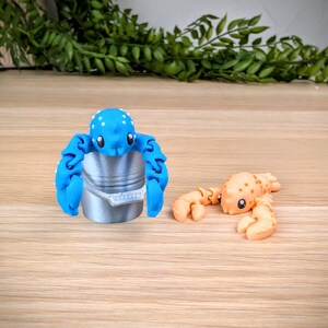 May include: Two small, colourful, 3D printed lobster figurines. One is blue with white spots and is holding a small silver bucket. The other is orange with white spots.
